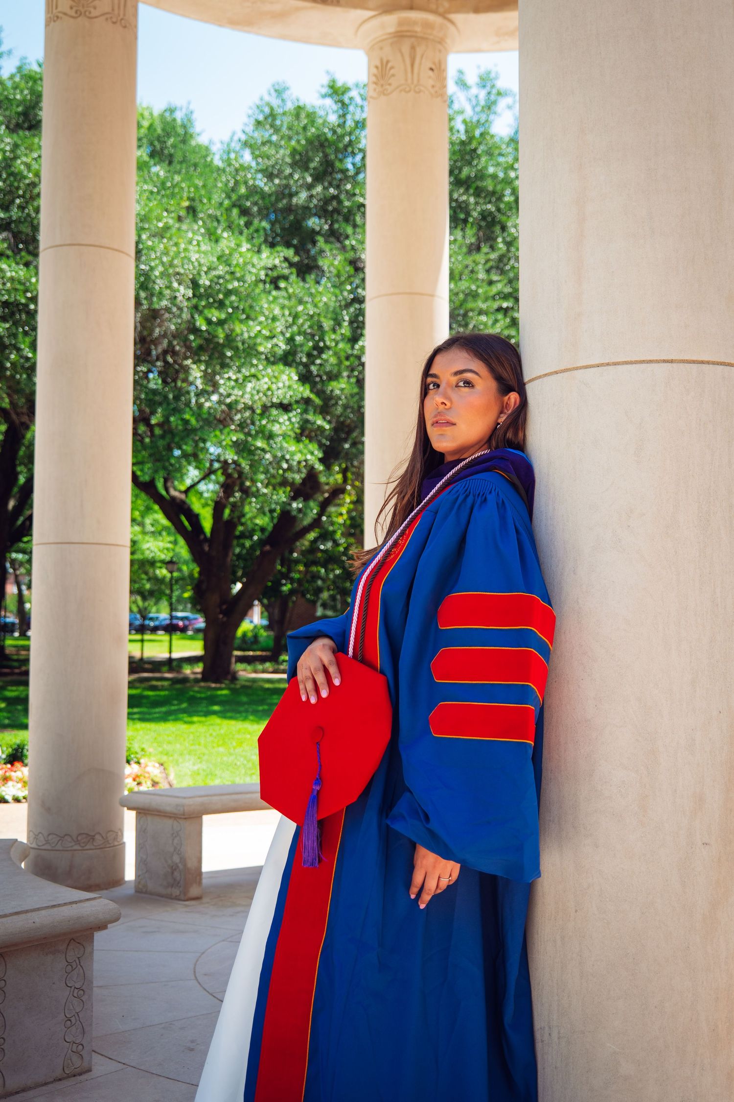 Female doctoral graduate in blue and red gown leans against column holding red cap in campus rotunda.