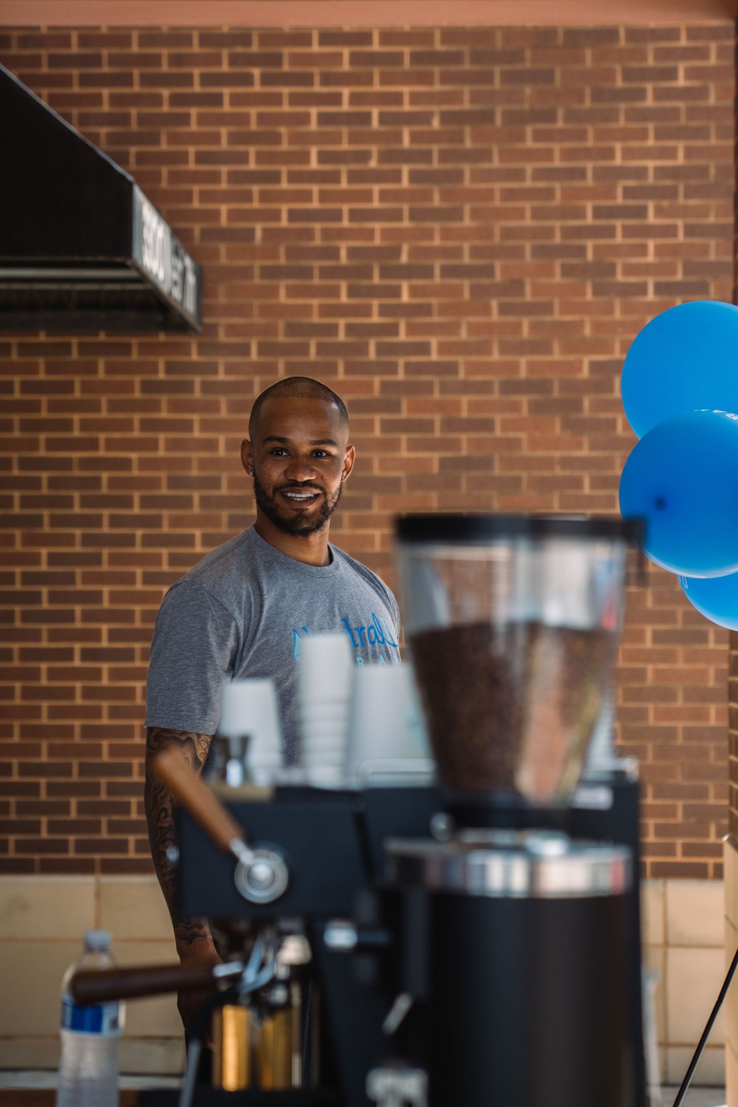 Smiling barista stands behind a coffee grinder with blue balloons and brick wall backdrop.