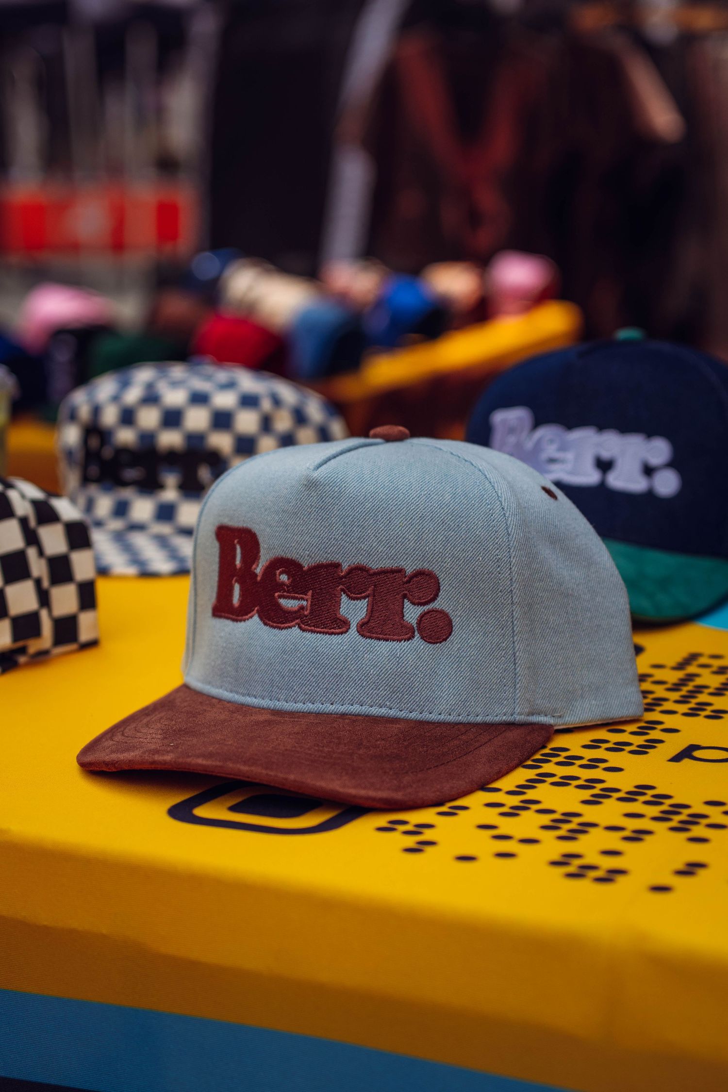 Gray and burgundy Berr. snapback cap displayed on a yellow table at a retail store with colorful hats in background.