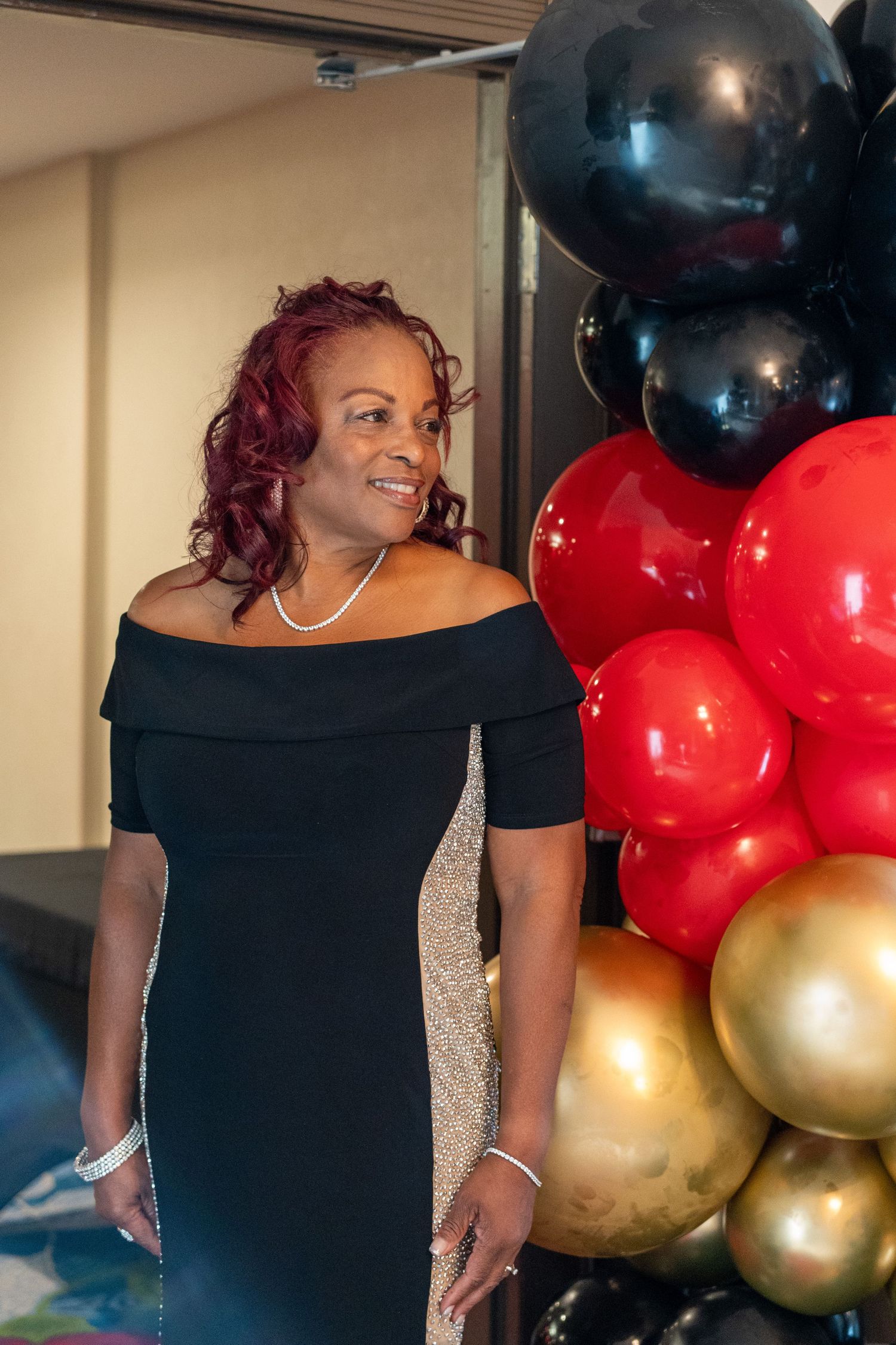 Woman in black off-shoulder dress smiling beside black, red, and gold balloon arrangement at elegant event.