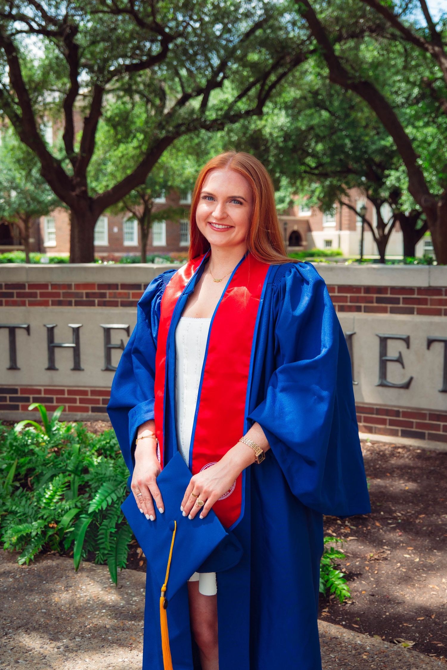 A smiling woman in blue graduation gown with red stole poses outdoors on a university campus.