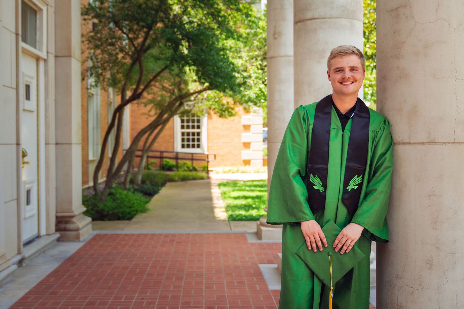 Young man in green graduation gown smiling outdoors near brick columns on a college campus.