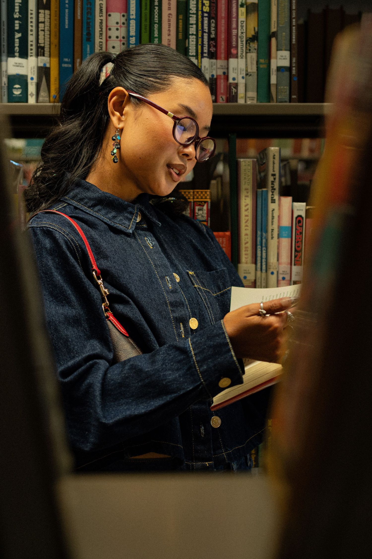 Young woman in denim jacket smiling while reading a book in a colorful library.