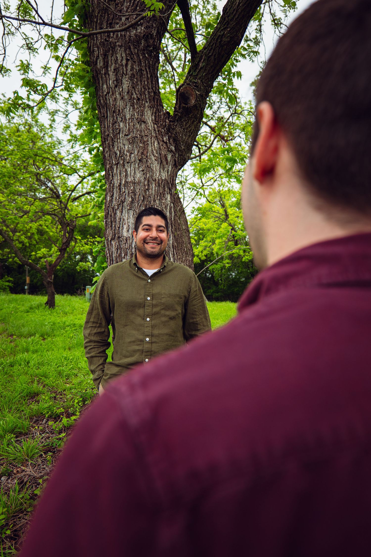 Two men in casual shirts smile at each other outdoors near a large tree in a lush green park.
