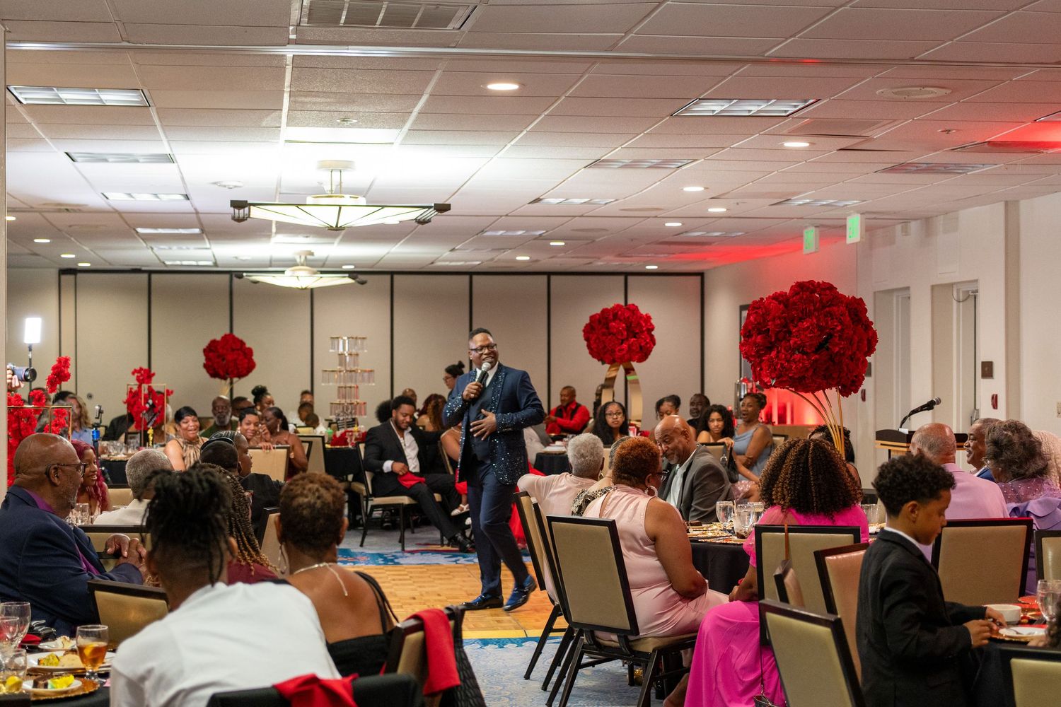 Elegant banquet event with guests seated at tables, a speaker at the podium, and red floral decorations in a ballroom.