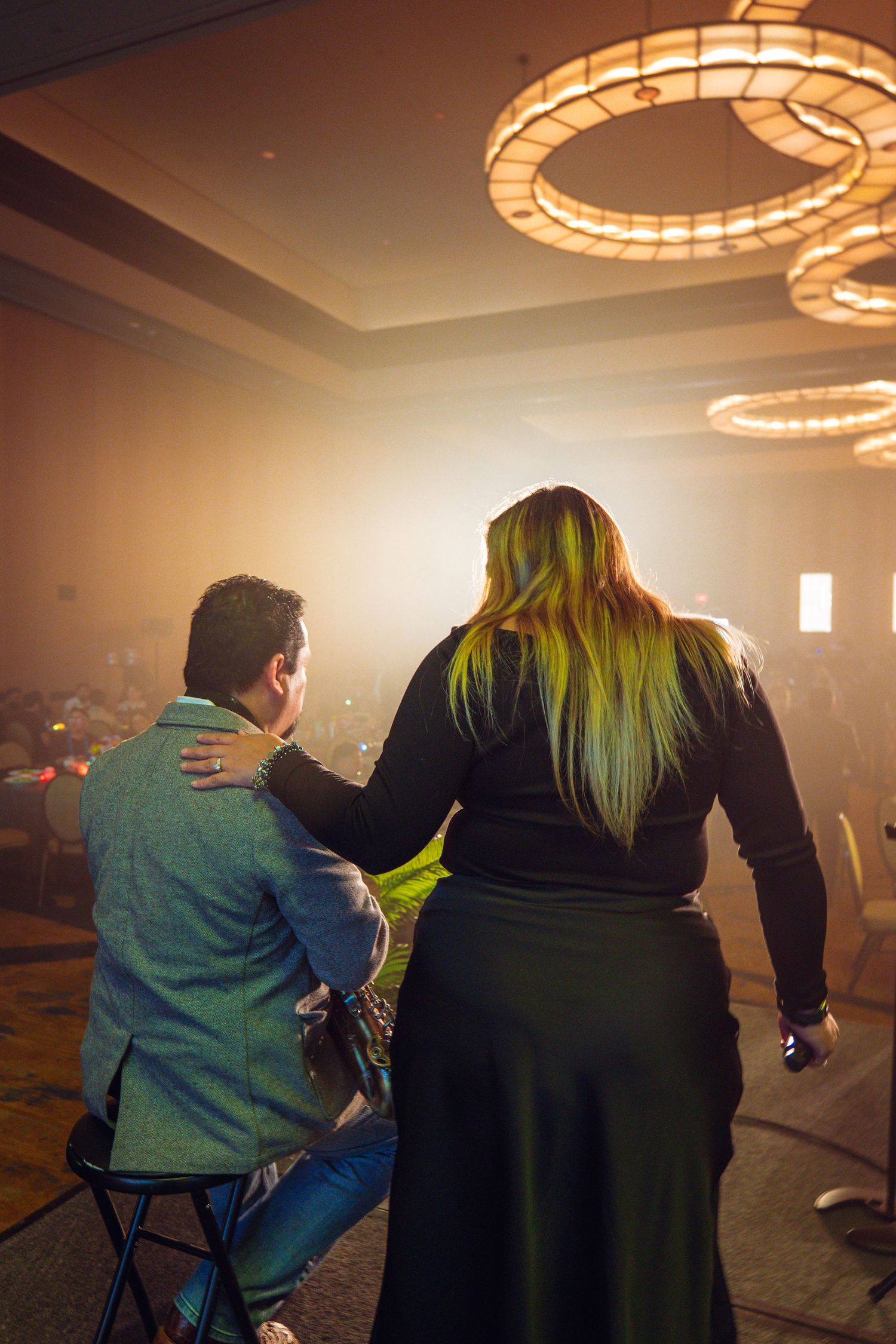 Two performers on stage in a warmly lit ballroom, woman with blonde hair in black dress beside seated man in gray jacket.