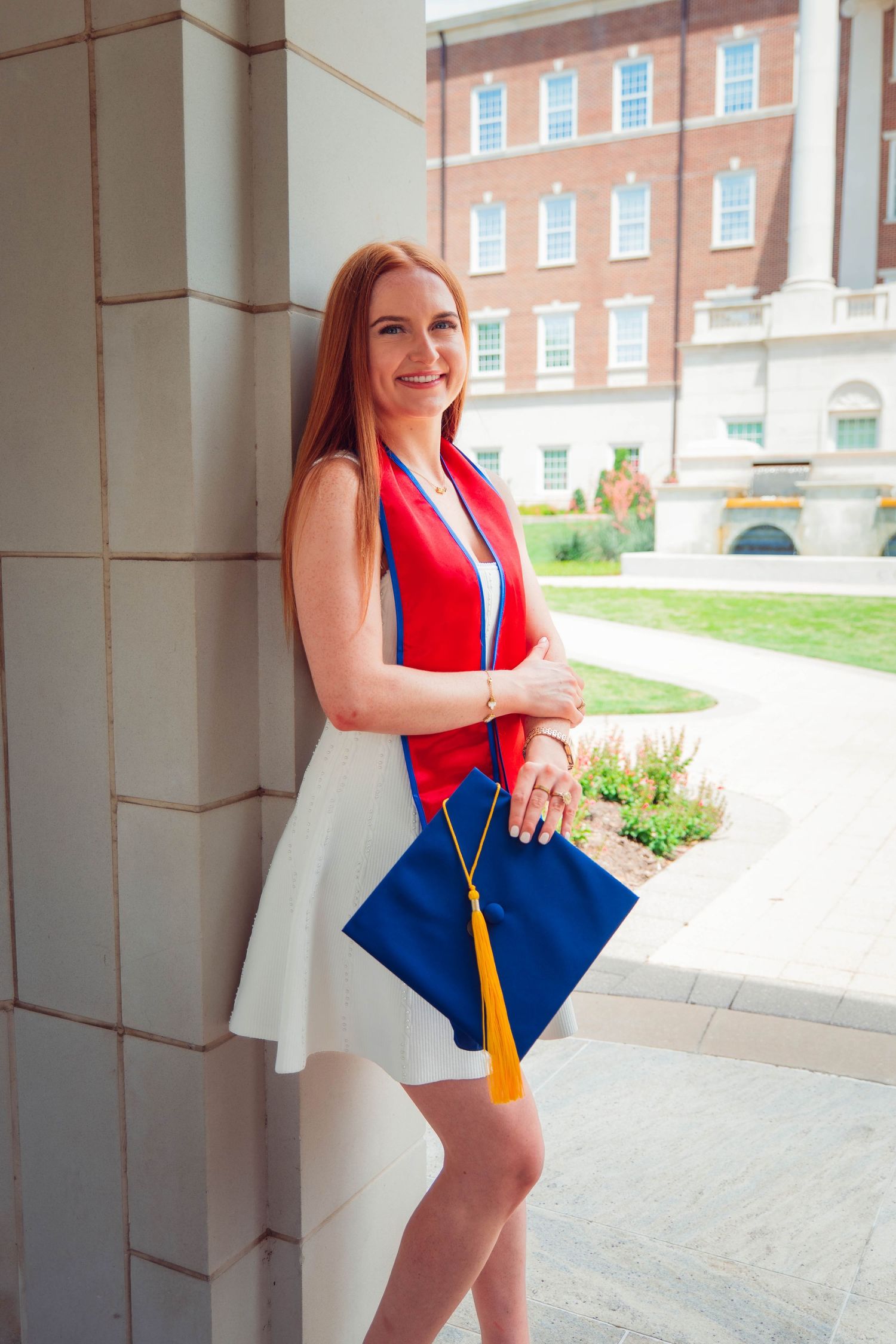 Smiling redhead woman in white dress holding blue graduation cap with yellow tassel, wearing red sash outdoors.