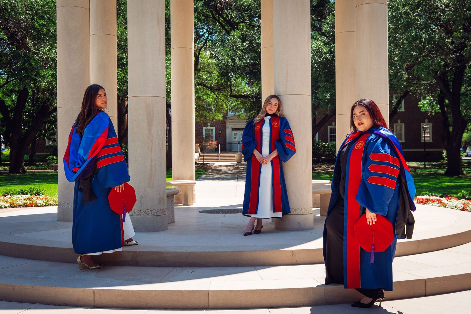 Three graduates in blue and red doctoral robes posing under a columned outdoor pavilion.