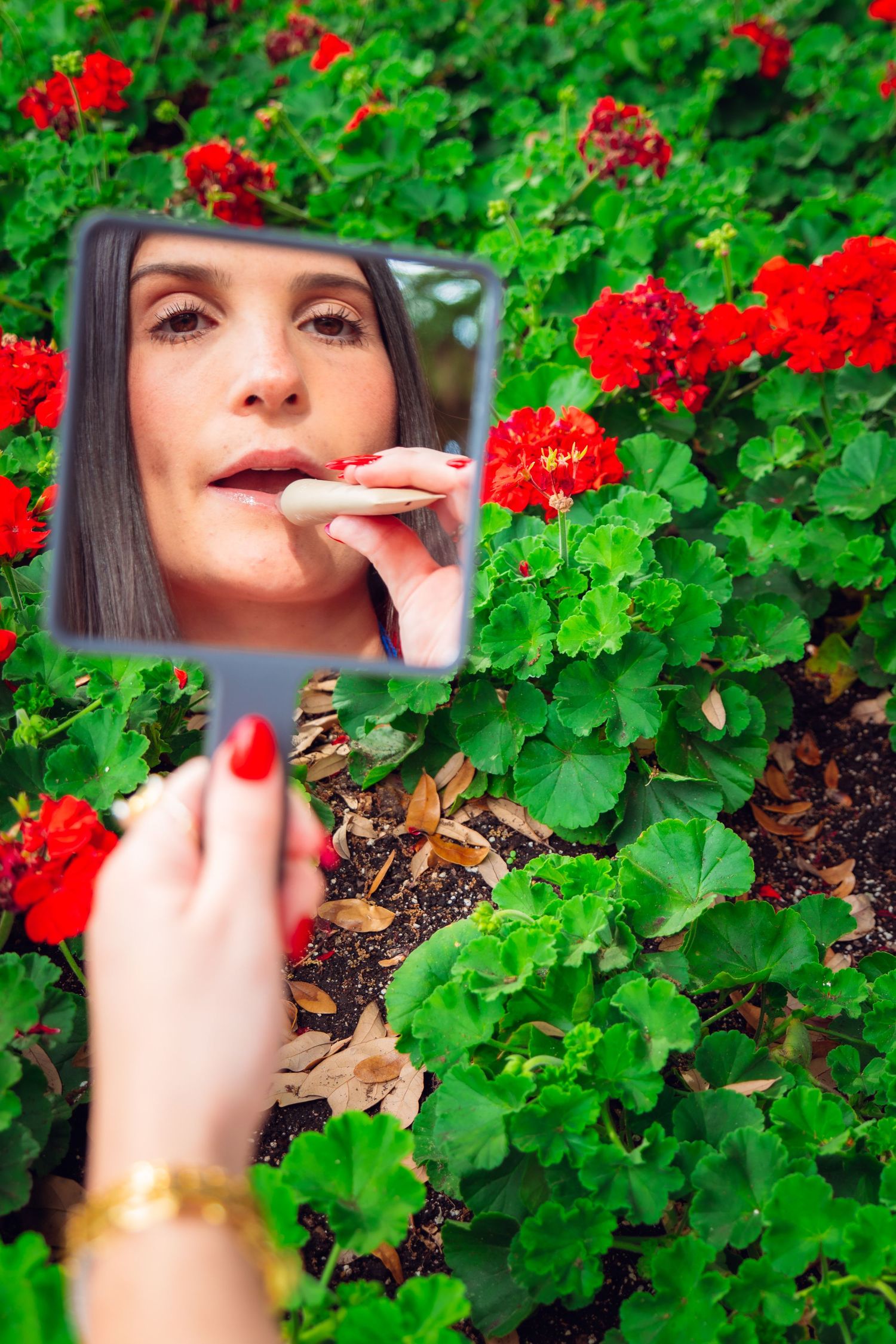 Woman checking her lipstick in a handheld mirror surrounded by vibrant red geranium flowers outdoors.