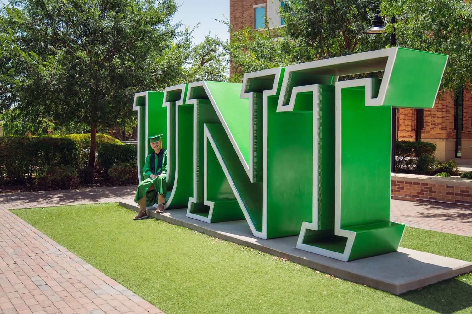 Graduate in cap and gown posing beside large green UNT letter sign on University of North Texas campus.