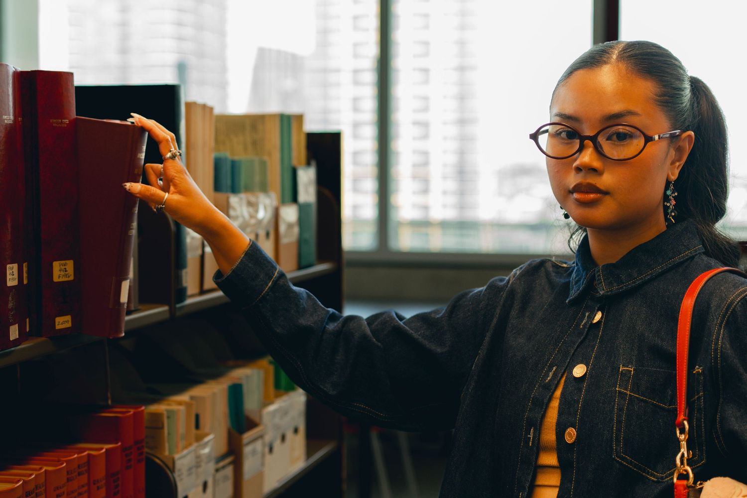 Young woman with glasses browsing books on a shelf in a modern library or bookstore.