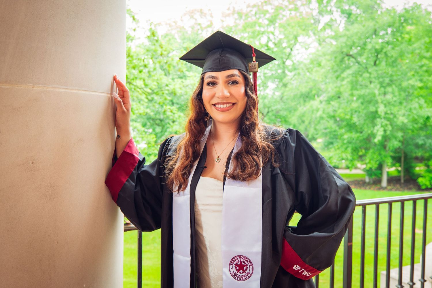 Young woman in black graduation cap and gown posing beside a stone column with green trees.