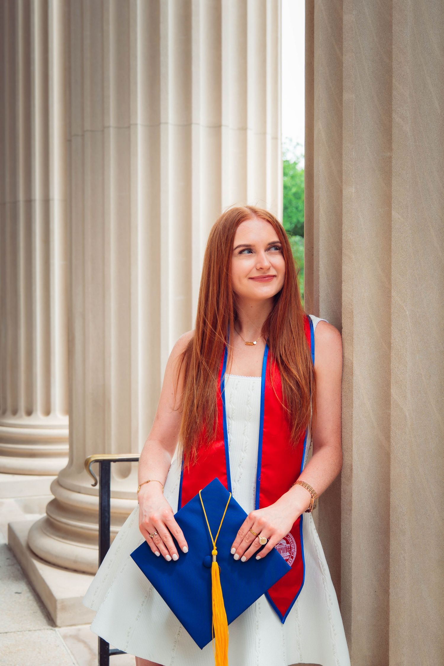 Young woman in white dress with red graduation stole holds blue cap beside stone columns, smiling upward.