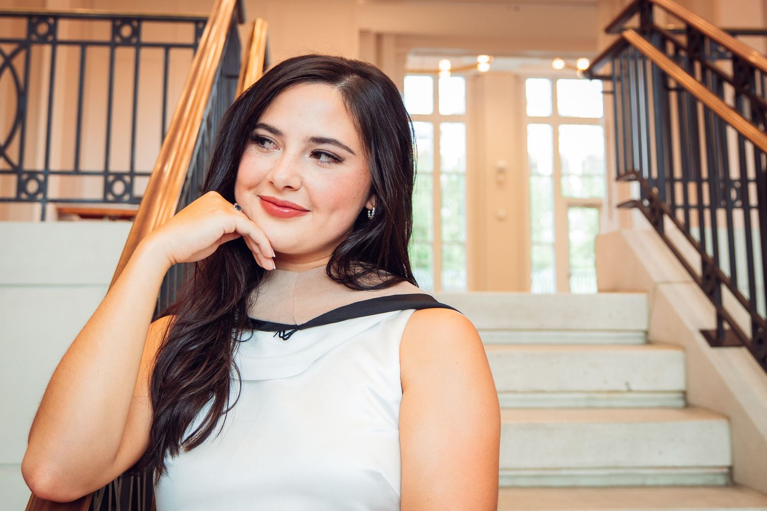 Smiling young woman in white dress posing on staircase in elegant indoor setting.