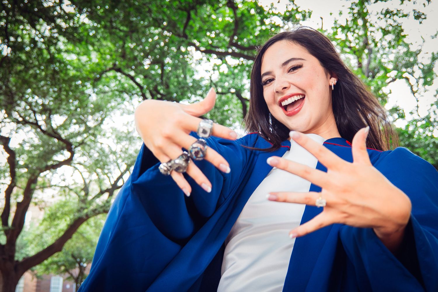 Excited young woman in blue graduation gown celebrating outdoors with trees in background.