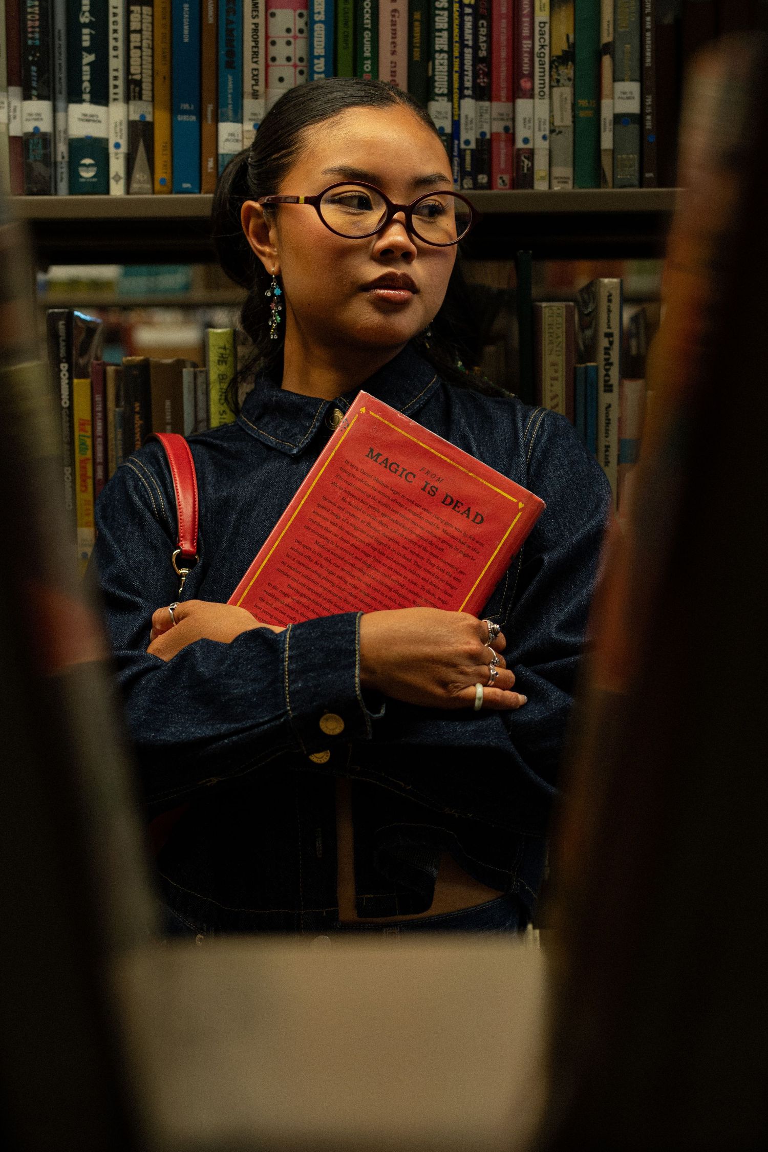 Young woman with glasses holding a red Magic is Dead book among library bookshelves.