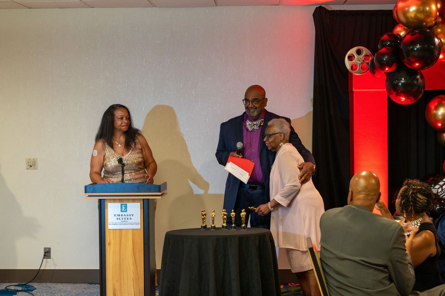 Award ceremony with three people at podium, red and black balloon decor, trophies on table at formal event.