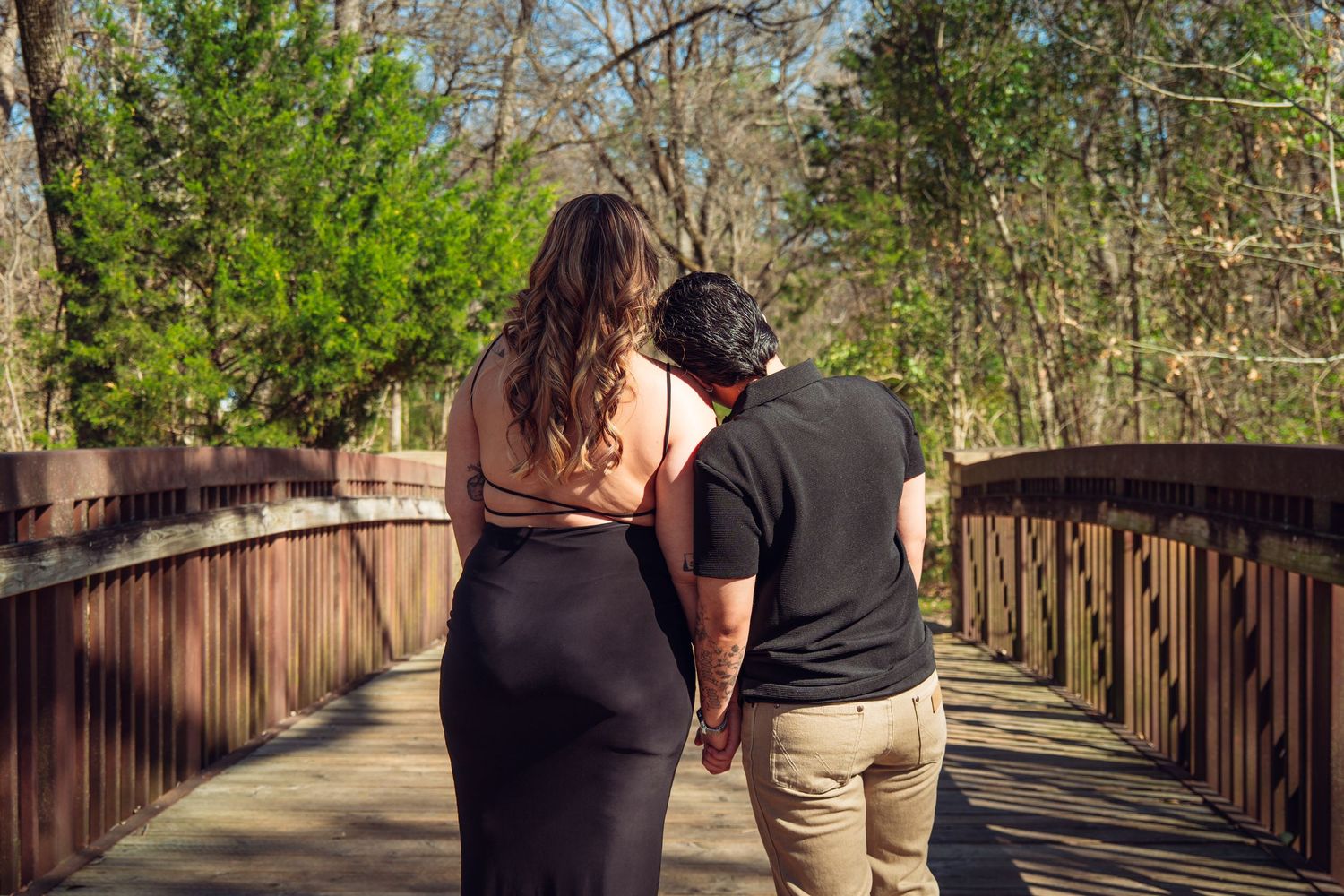 Couple holding hands walking on a wooden bridge surrounded by lush green trees in a park.