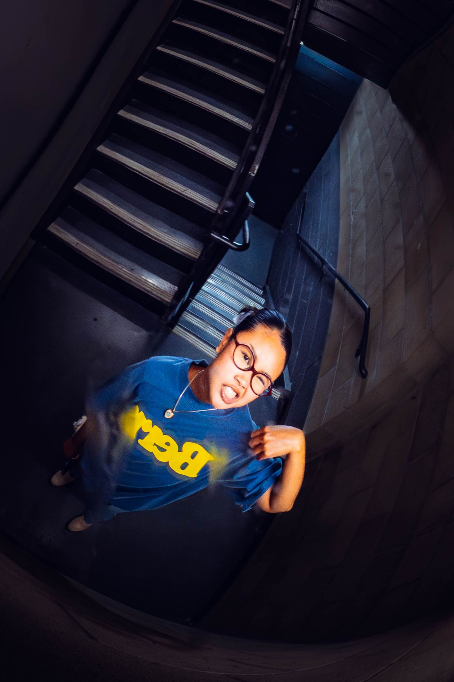Young woman in blue graphic tee and glasses stands at bottom of dark spiral staircase, looking up at camera.
