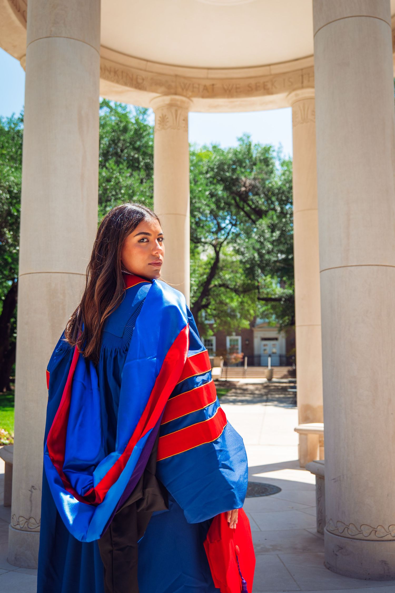 Female doctoral graduate in blue and red regalia posing between stone columns.