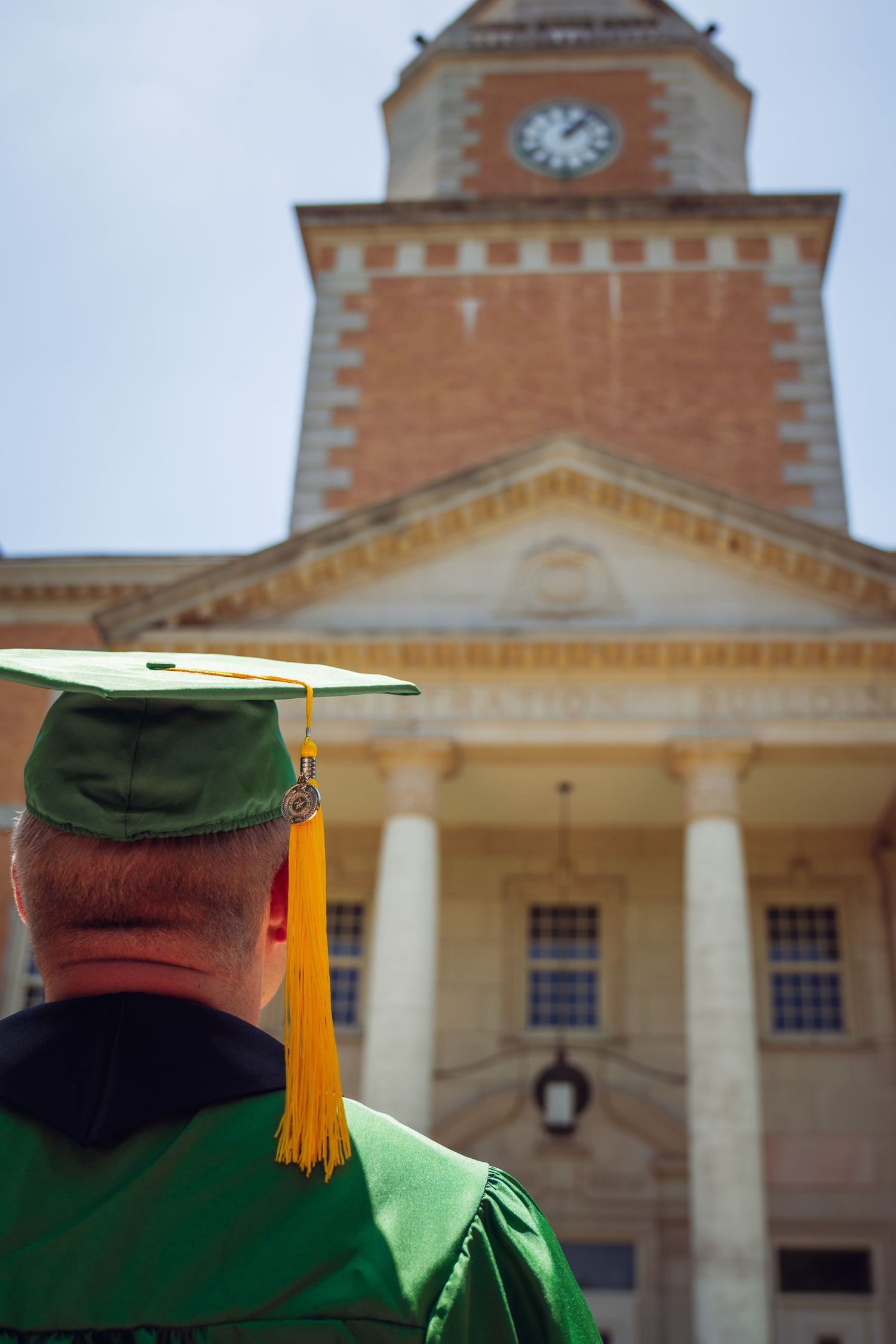Graduate in green cap and gown stands before a historic brick clock tower building at commencement ceremony.