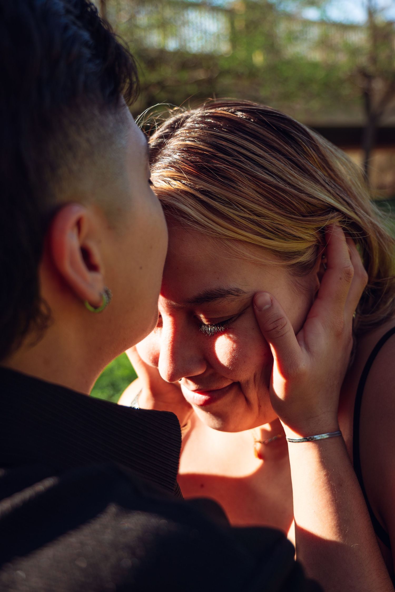 A tender moment between two people, one gently cupping the other's smiling face in warm golden sunlight outdoors.