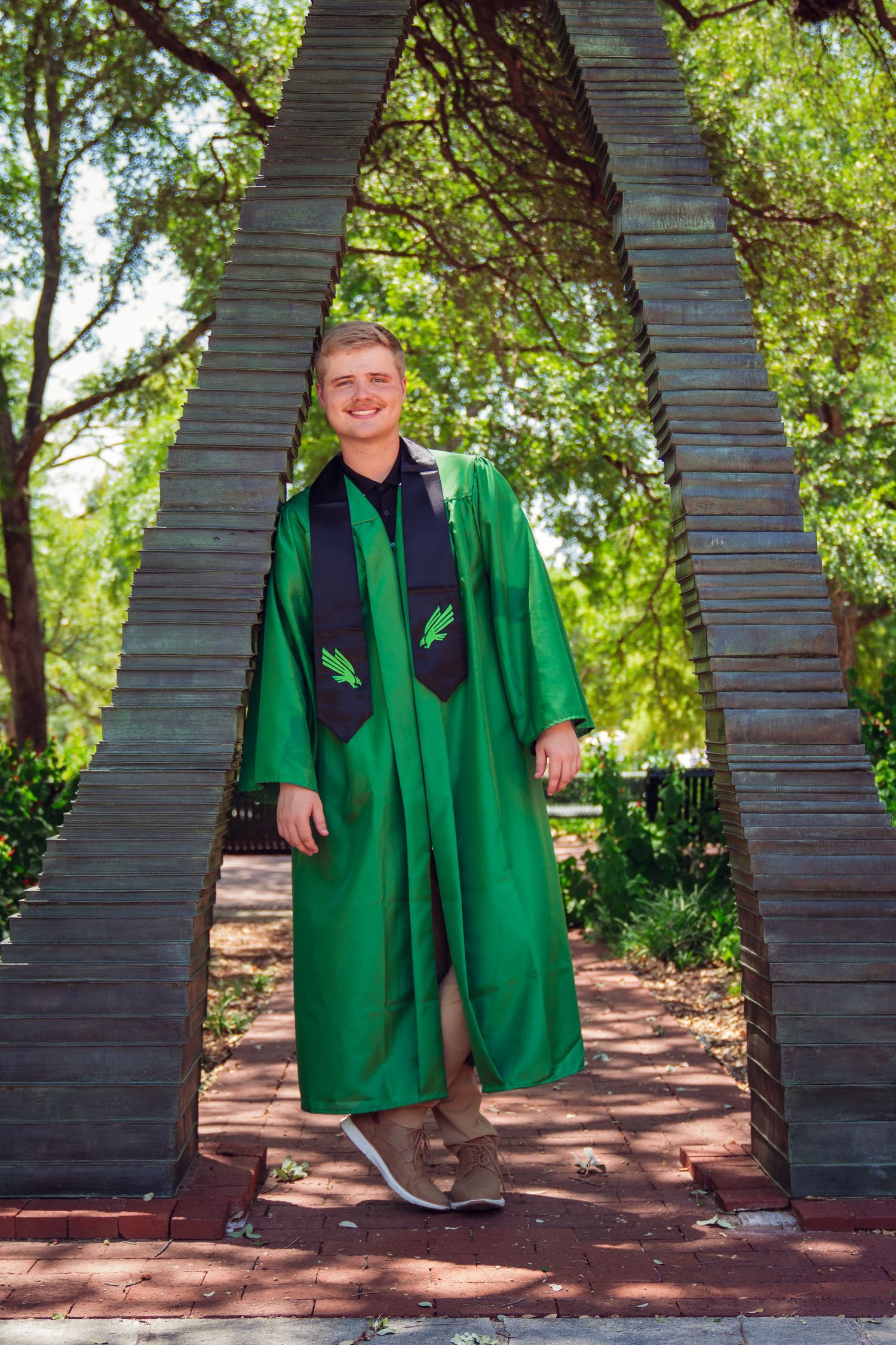 Smiling graduate in green cap and gown poses under an arch monument surrounded by lush trees on a brick pathway.