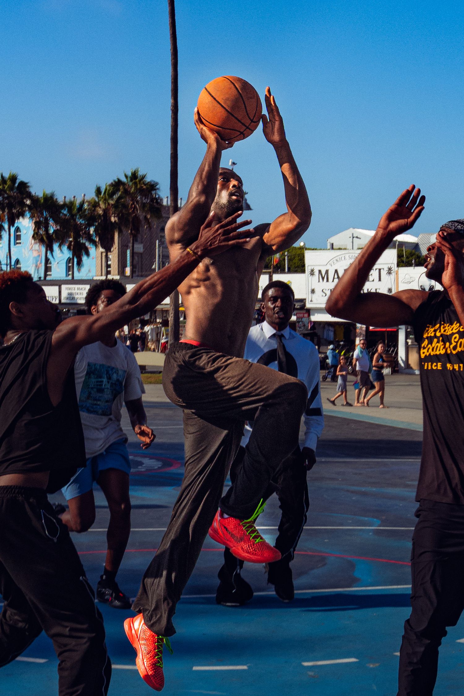 Shirtless basketball player jumps to shoot over defenders on an outdoor court at Venice Beach, CA.