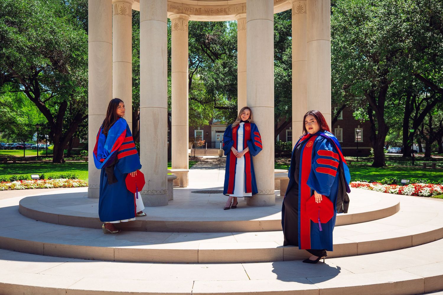 Three graduates in blue and red doctoral robes pose at an outdoor columned rotunda on a sunny university campus.