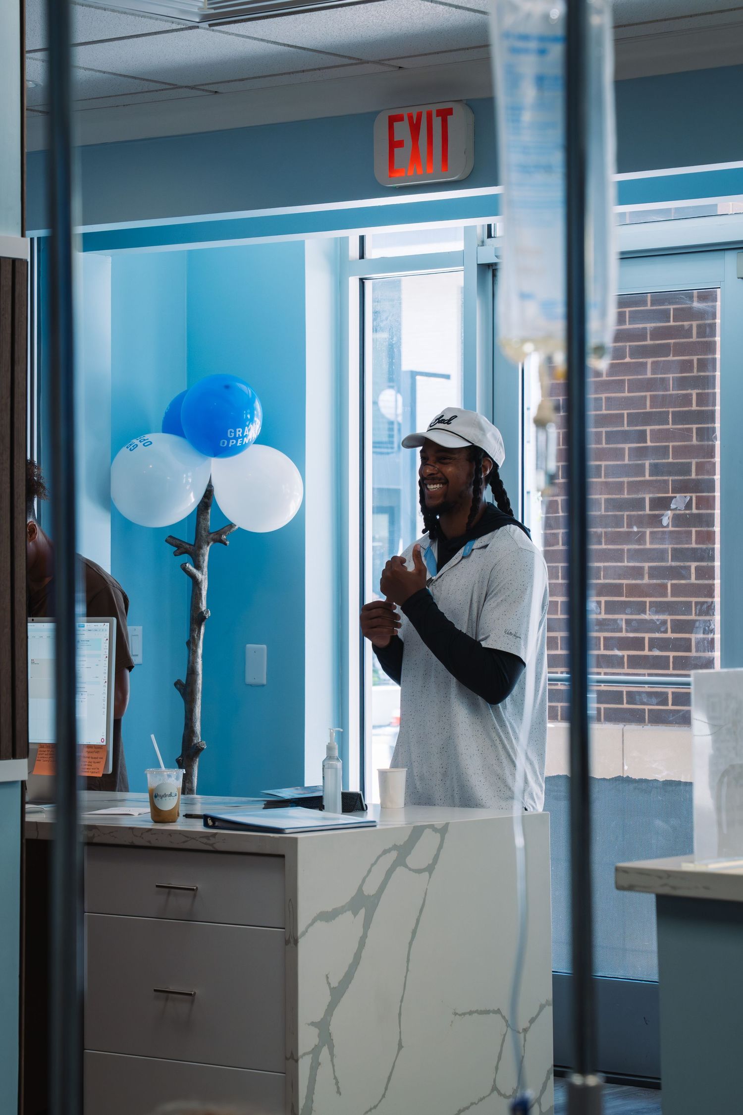 Man in white cap smiling at office reception desk with blue and white balloons near an exit sign.