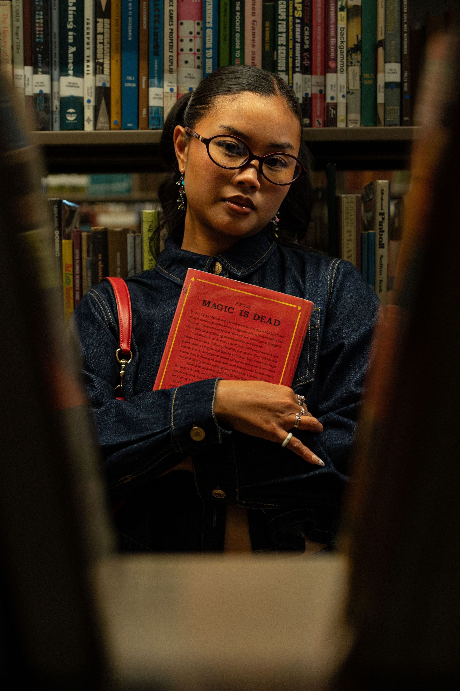Young woman in denim jacket holding a red book titled Magic is Dead in a library.
