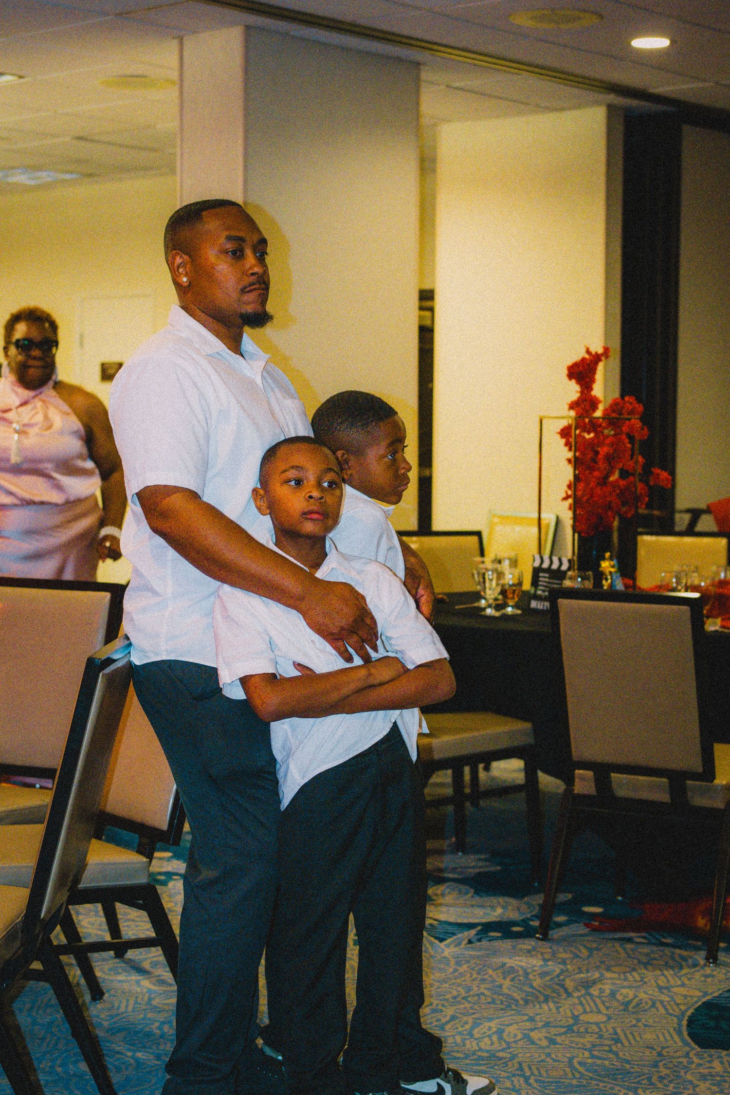 A man and two young boys dressed in white shirts stand together at a formal indoor event.