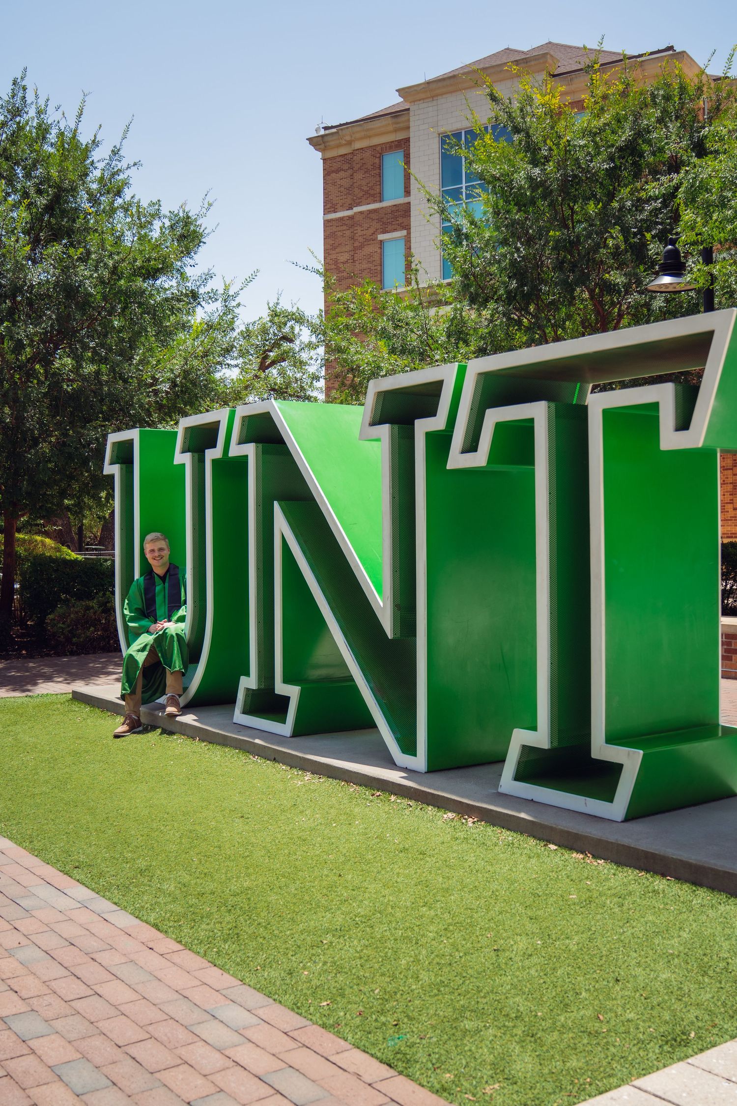 Person sitting in front of large green UNT letters sign on University of North Texas campus on a sunny day.