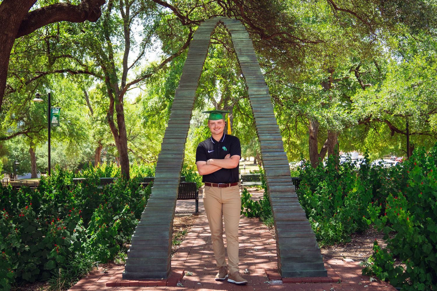 Graduate in cap poses under a large stone arch sculpture surrounded by lush green trees on a sunny campus.