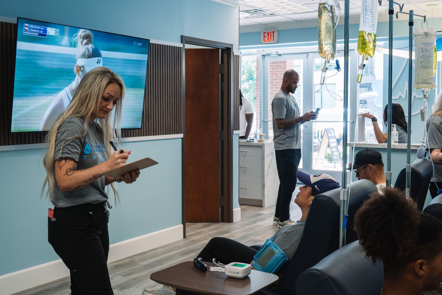 Medical staff attend to patients receiving IV drip treatments in a modern wellness infusion clinic with a TV on the wall.