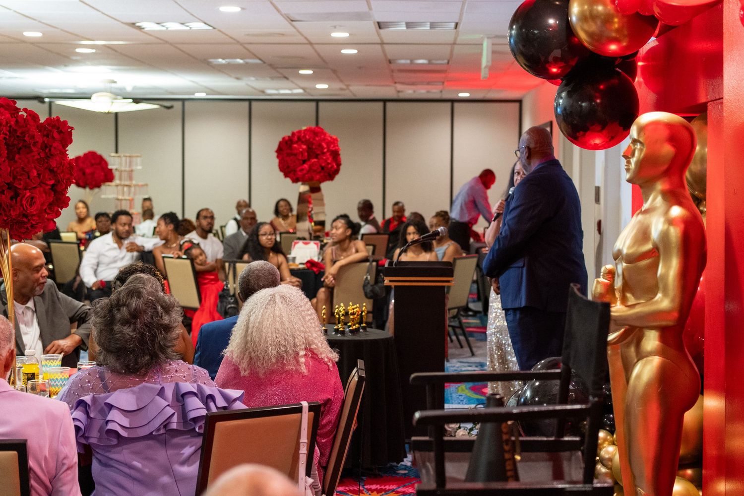 Elegant awards banquet with red and black decor, balloons, Oscar statue, and speaker at podium addressing seated guests.