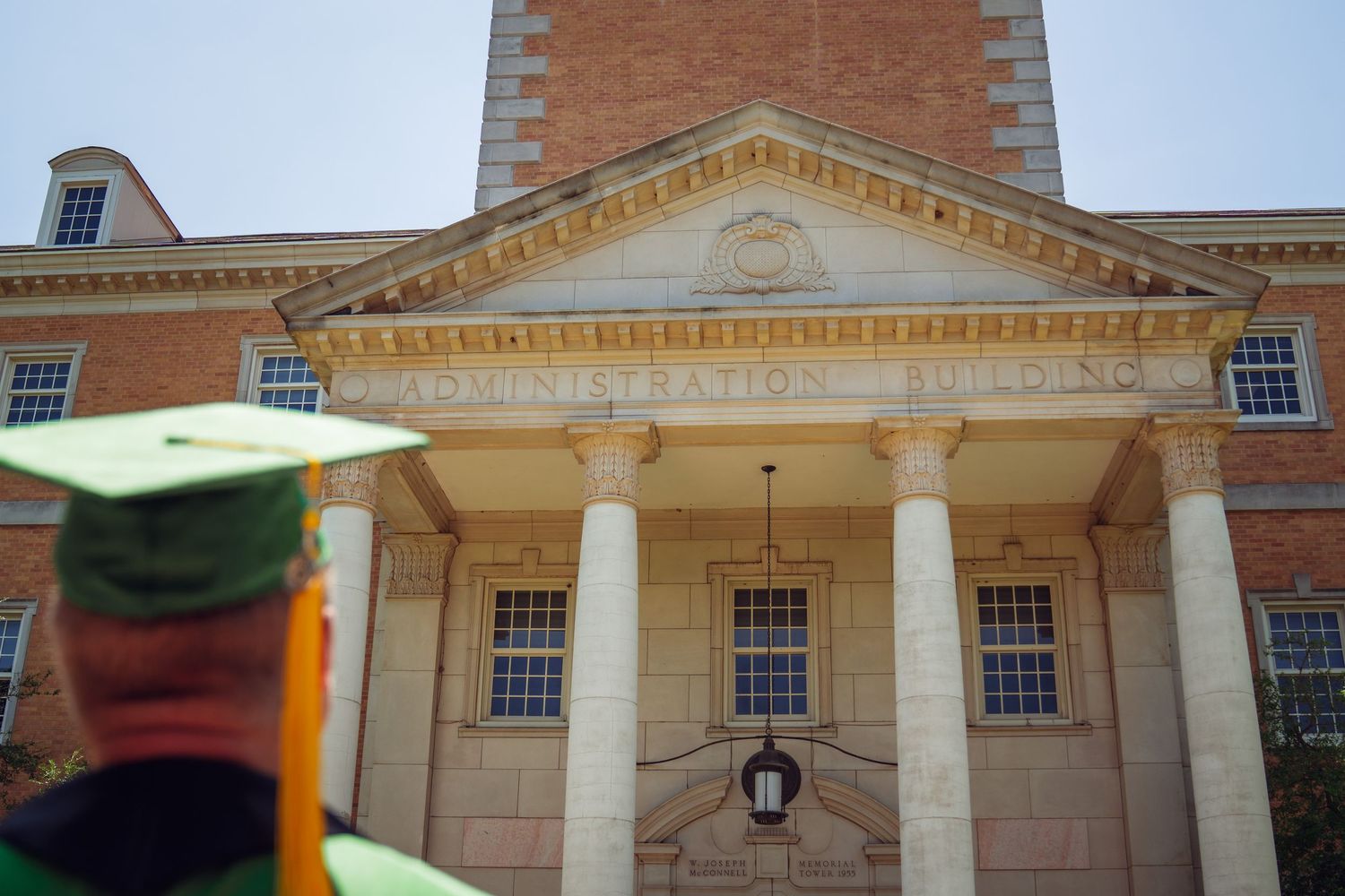 Graduate in cap and gown stands before a classic brick university administration building with white columns.
