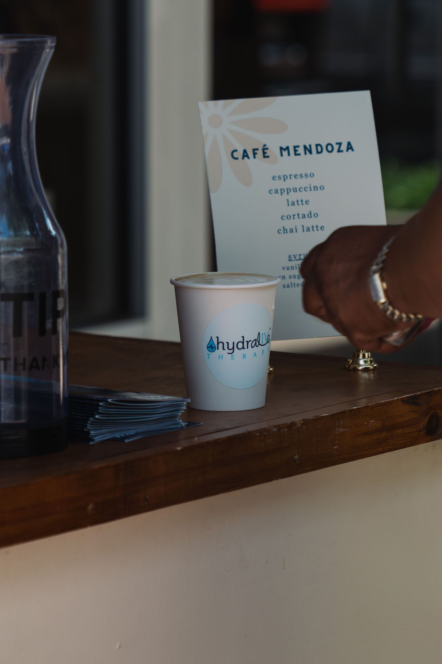 A hydralyte cup sits on a wooden counter at Café Mendoza beside a menu listing espresso, cappuccino, latte, cortado, and chai latte.