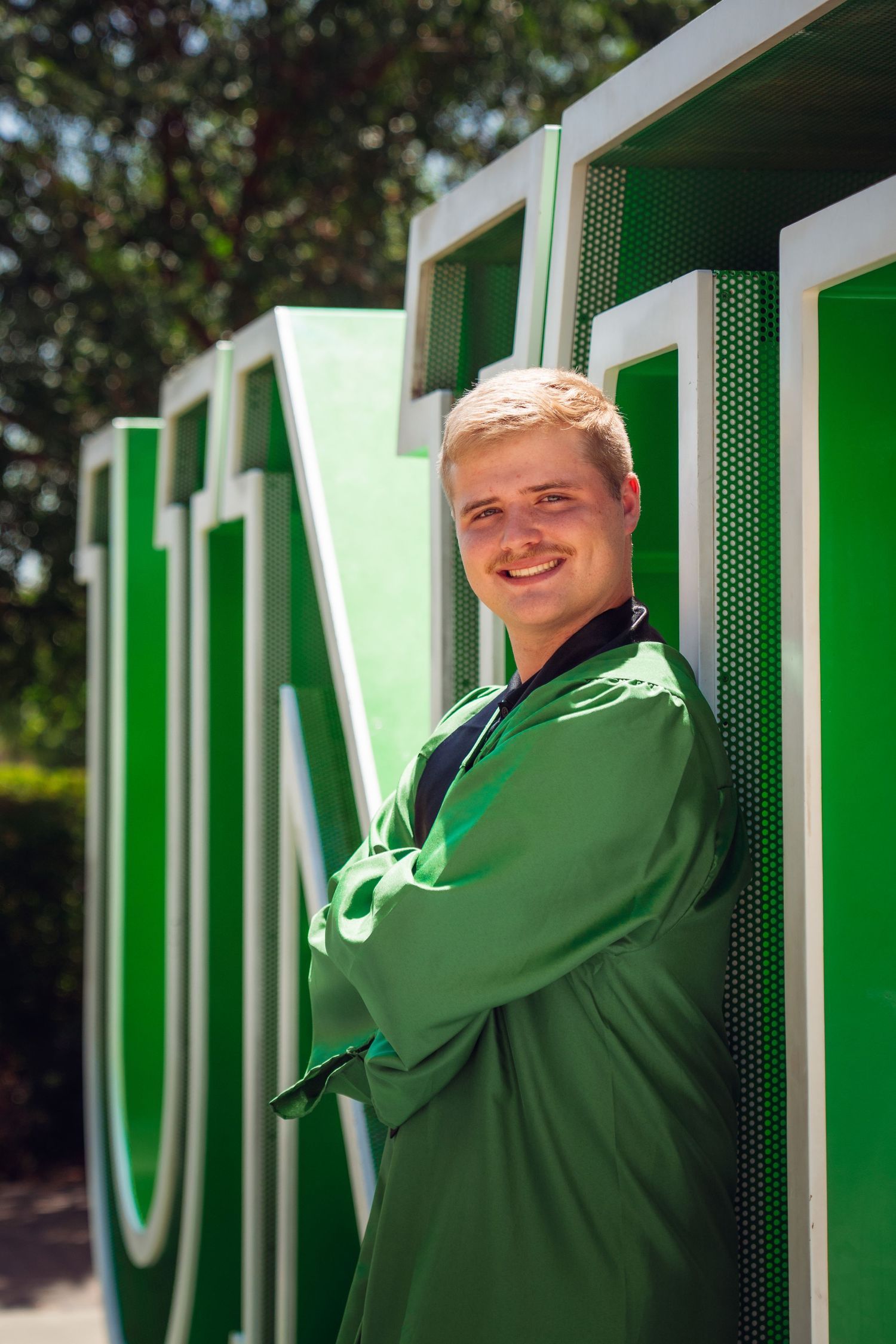 Smiling young man in green graduation gown stands with arms crossed beside large green university letters outdoors.