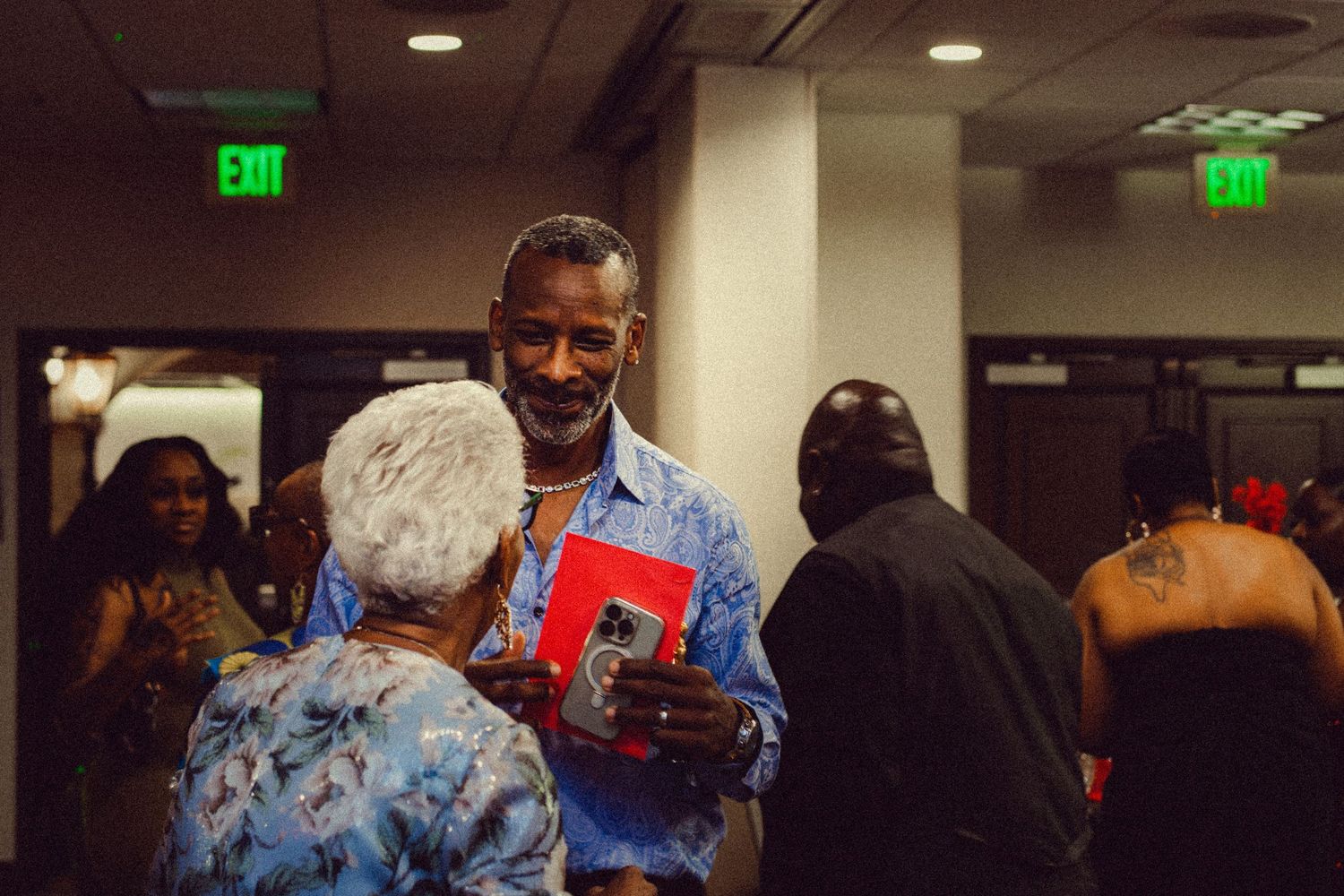 Man in blue floral shirt smiling and holding a book while conversing at an indoor social gathering event.