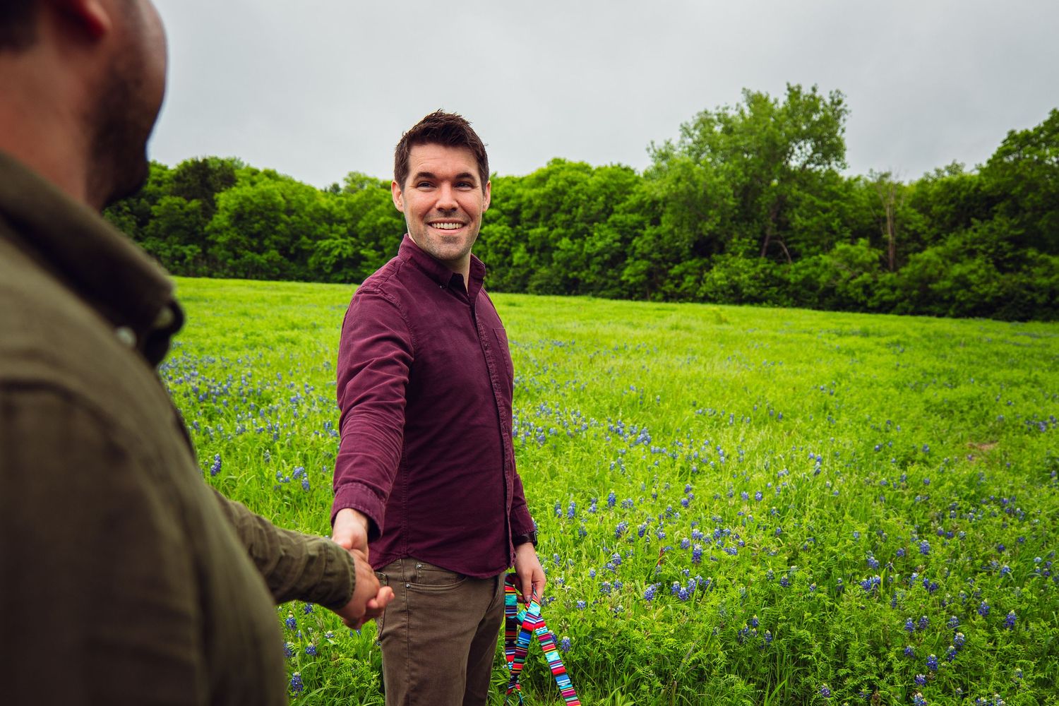 Two men holding hands in a field of bluebonnets, one smiling at camera.