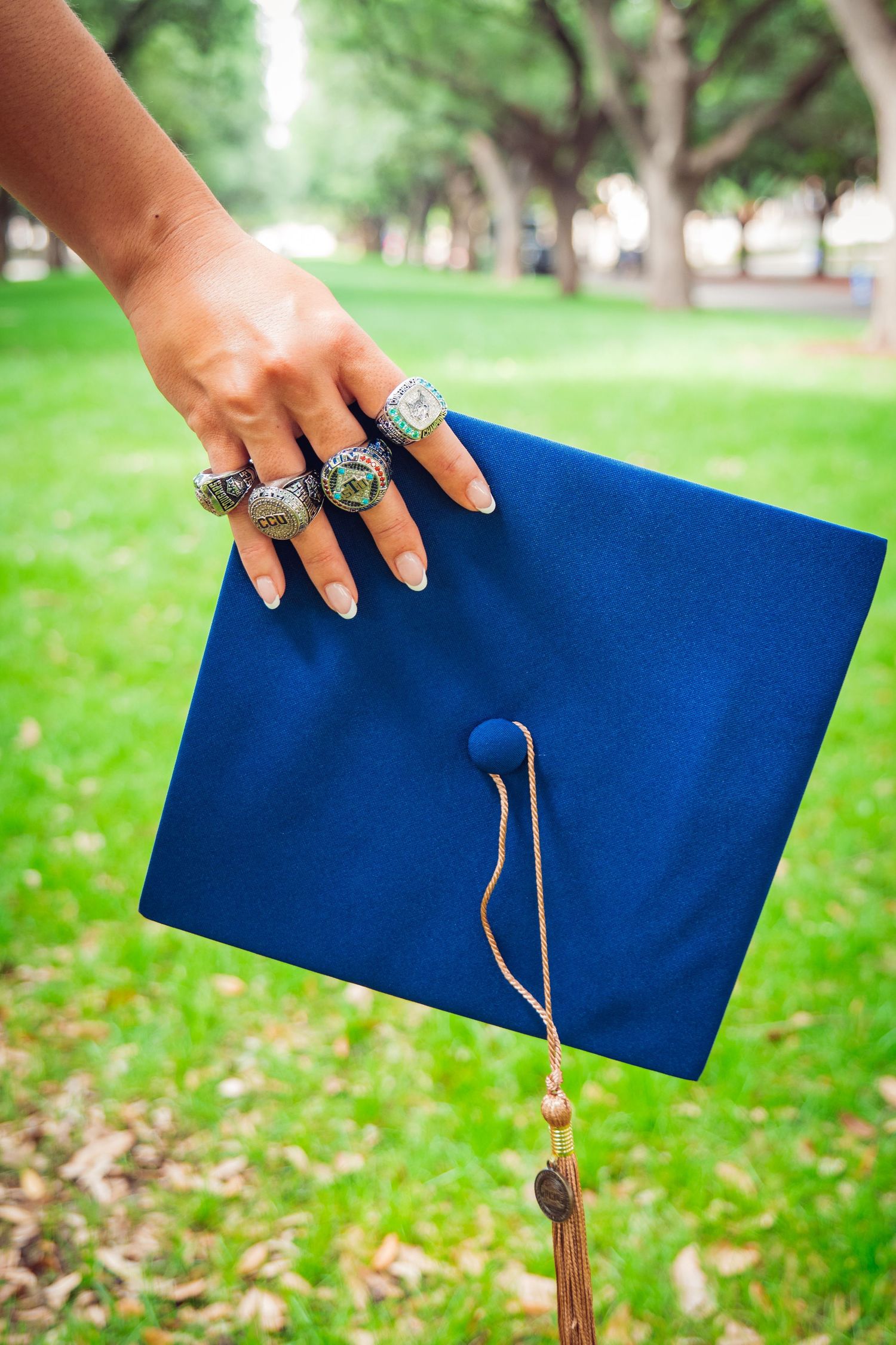 Hand adorned with championship rings holds blue graduation cap with gold tassel on green campus lawn.