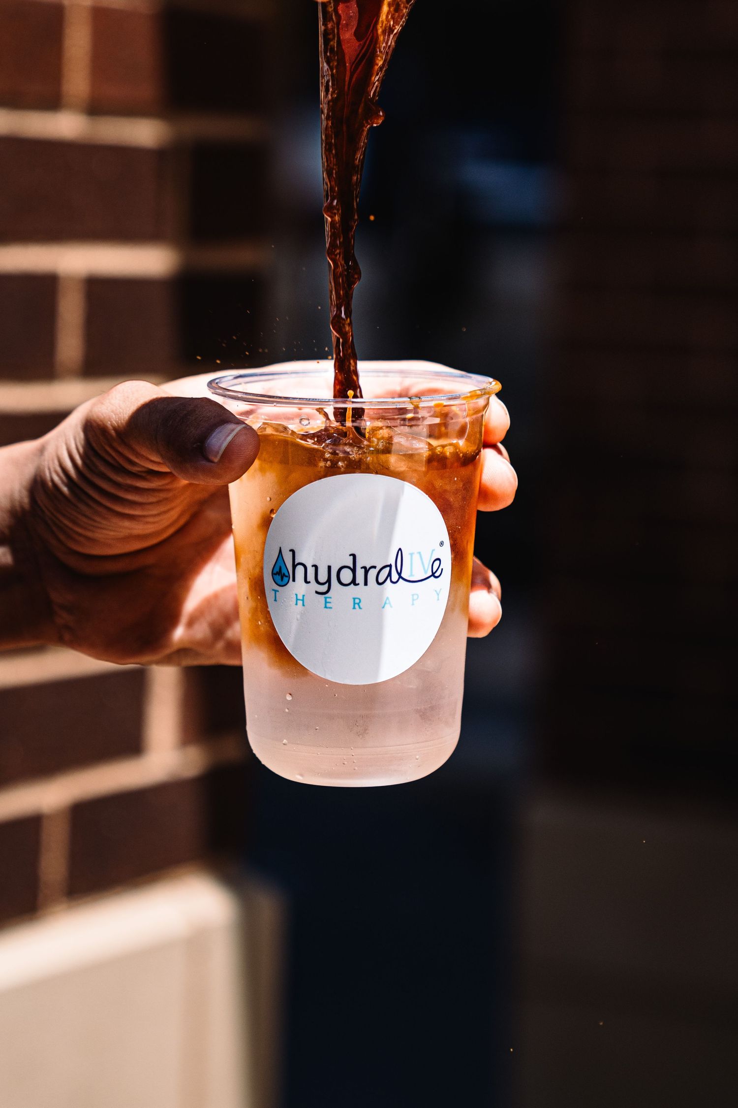 A hand holds a Hydrale Therapy cup being filled with iced tea outdoors near a brick wall.
