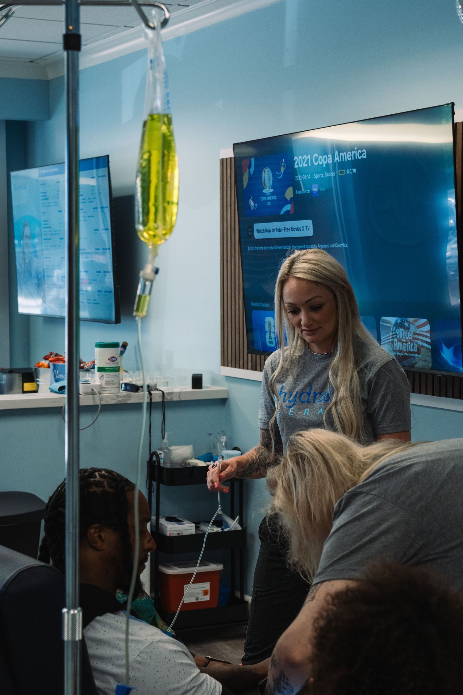 Healthcare worker administering IV drip therapy to patients in a modern medical clinic with TV screens in background.