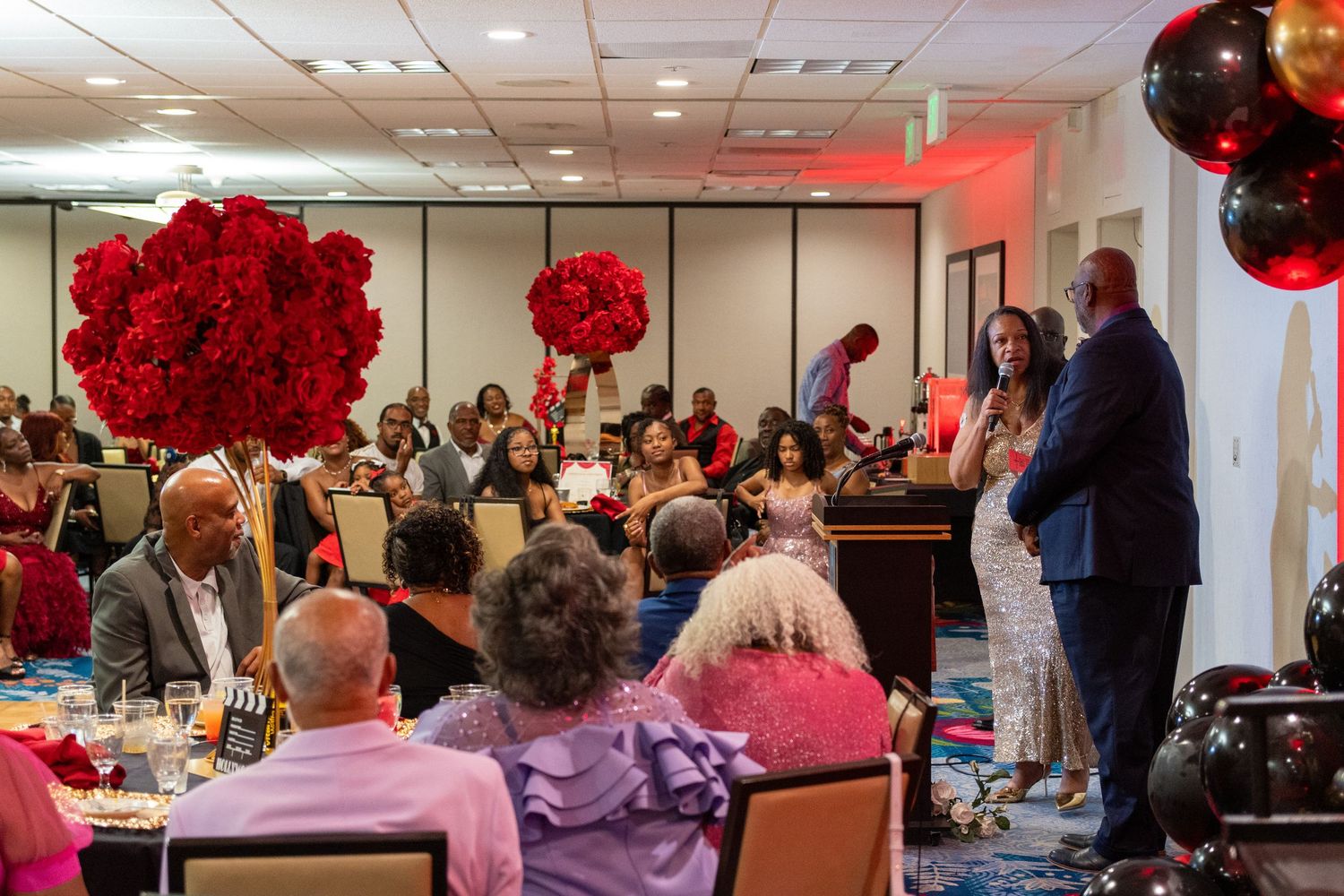 Elegant banquet hall with red rose centerpieces, black and gold balloons, and guests seated at tables during a formal event.