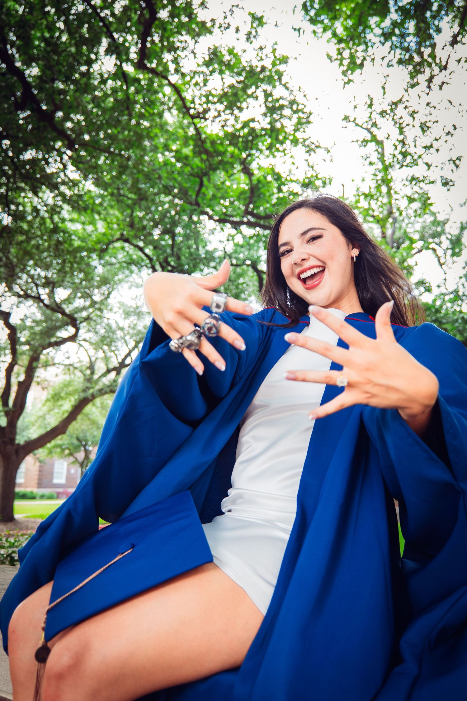 Excited female graduate in blue cap and gown showing rings while sitting outdoors.