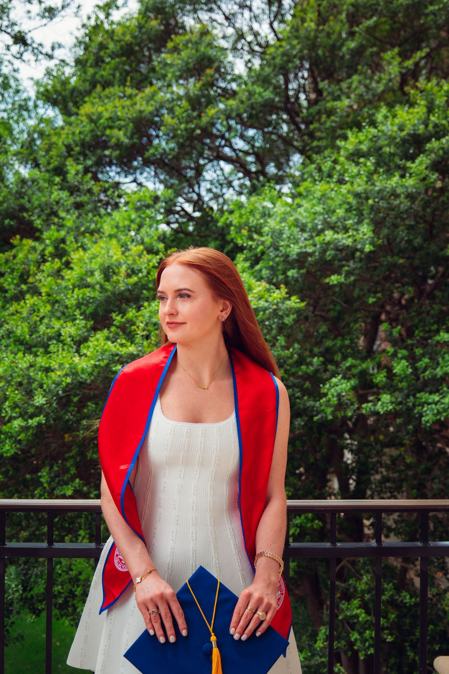 Red-haired woman in white dress with red and blue graduation regalia stands on balcony amid green trees.