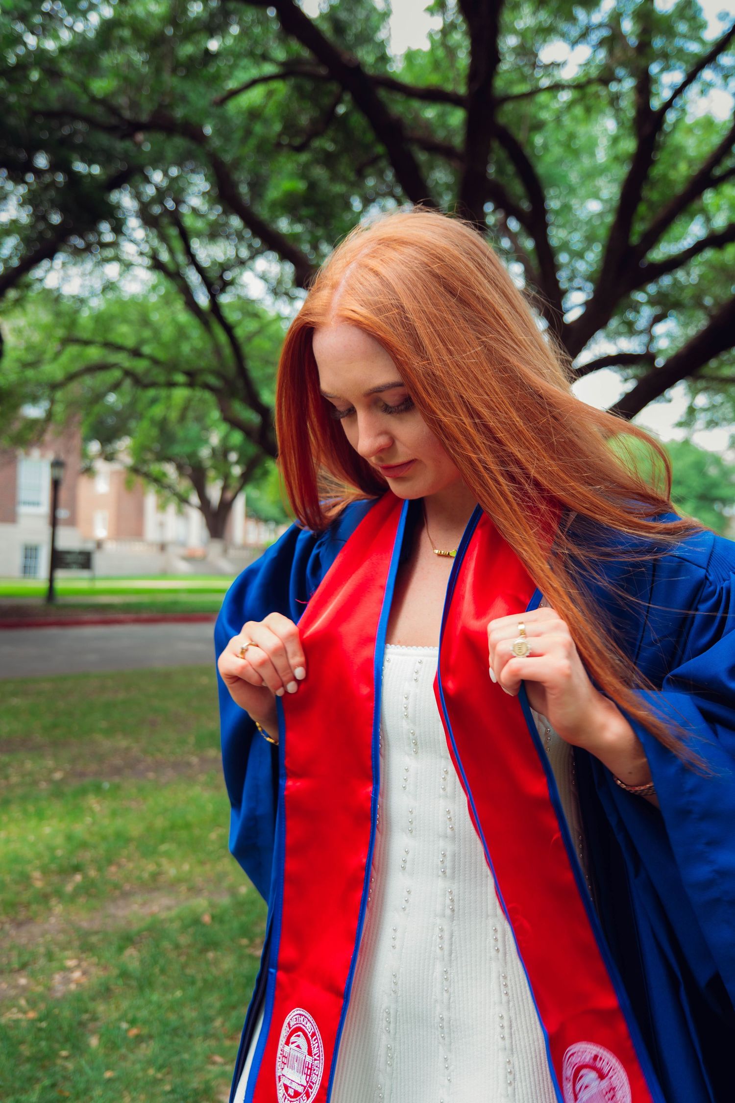 A young woman with long red hair wearing a blue graduation gown and red stole stands outdoors on a campus.