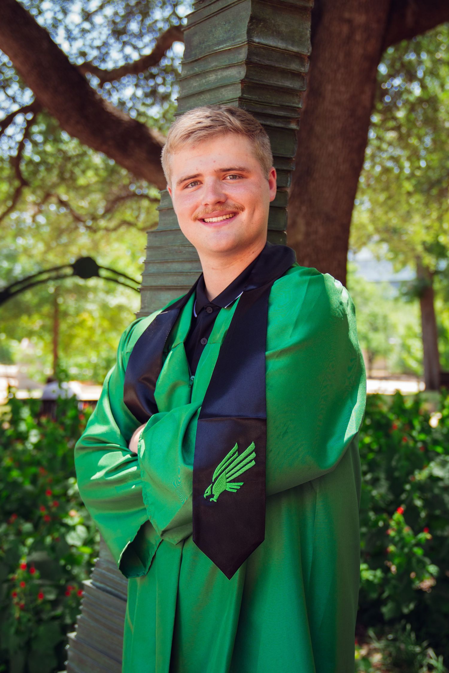 Young man smiling in green graduation gown with UNT stole, arms crossed, posing outdoors near tree.