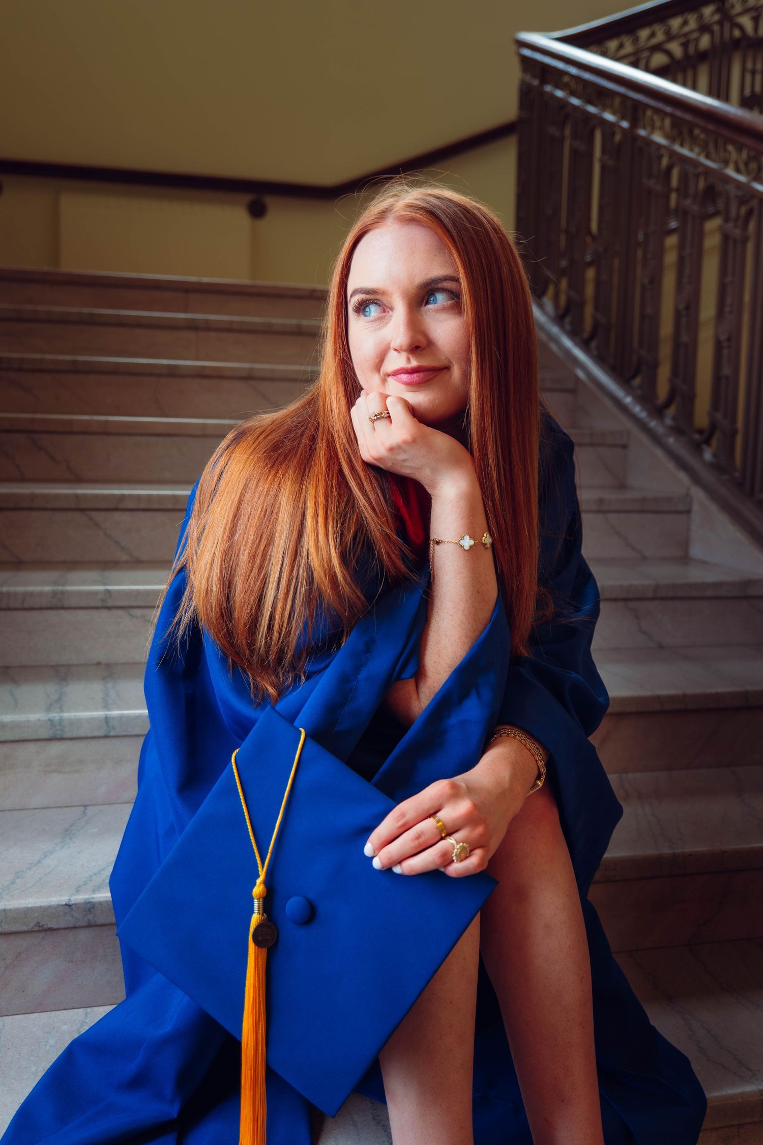 Red-haired graduate in blue cap and gown sits thoughtfully on marble stairs holding her mortarboard.